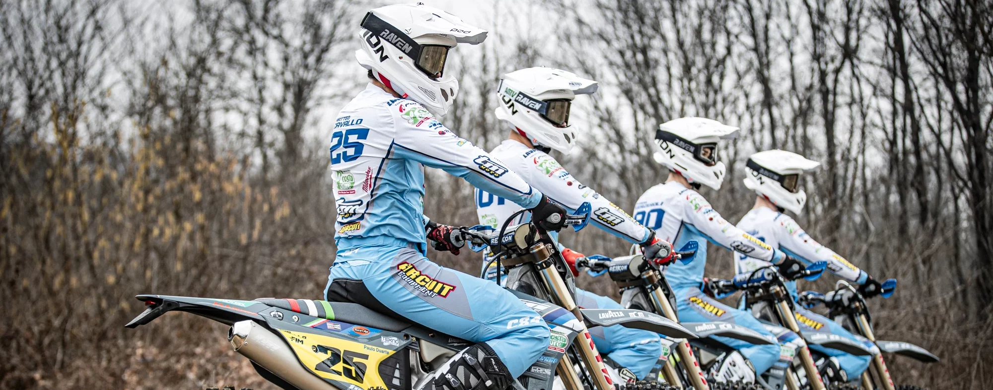 Vier motorcrossers in blauwe pakken en witte helmen op een rij in een kale bosomgeving.