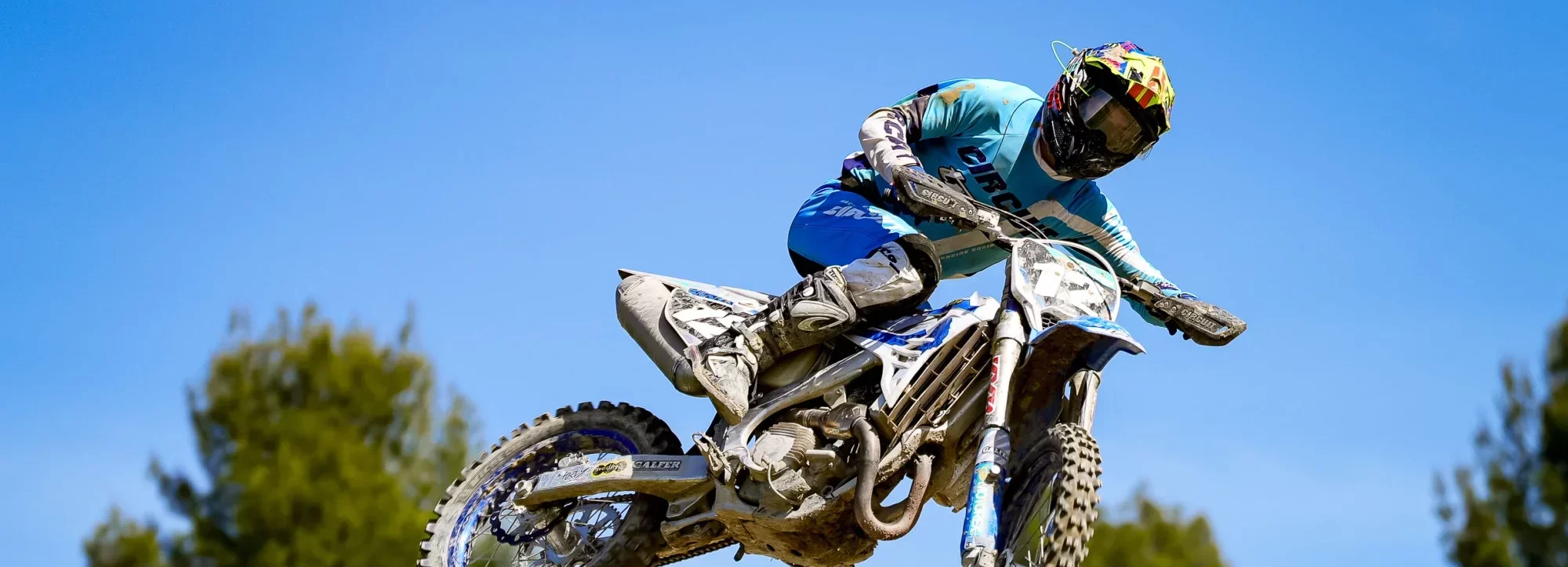 Motorcrosser maakt een hoge sprong boven een heuvelachtig terrein met bomen op de achtergrond onder een blauwe lucht.