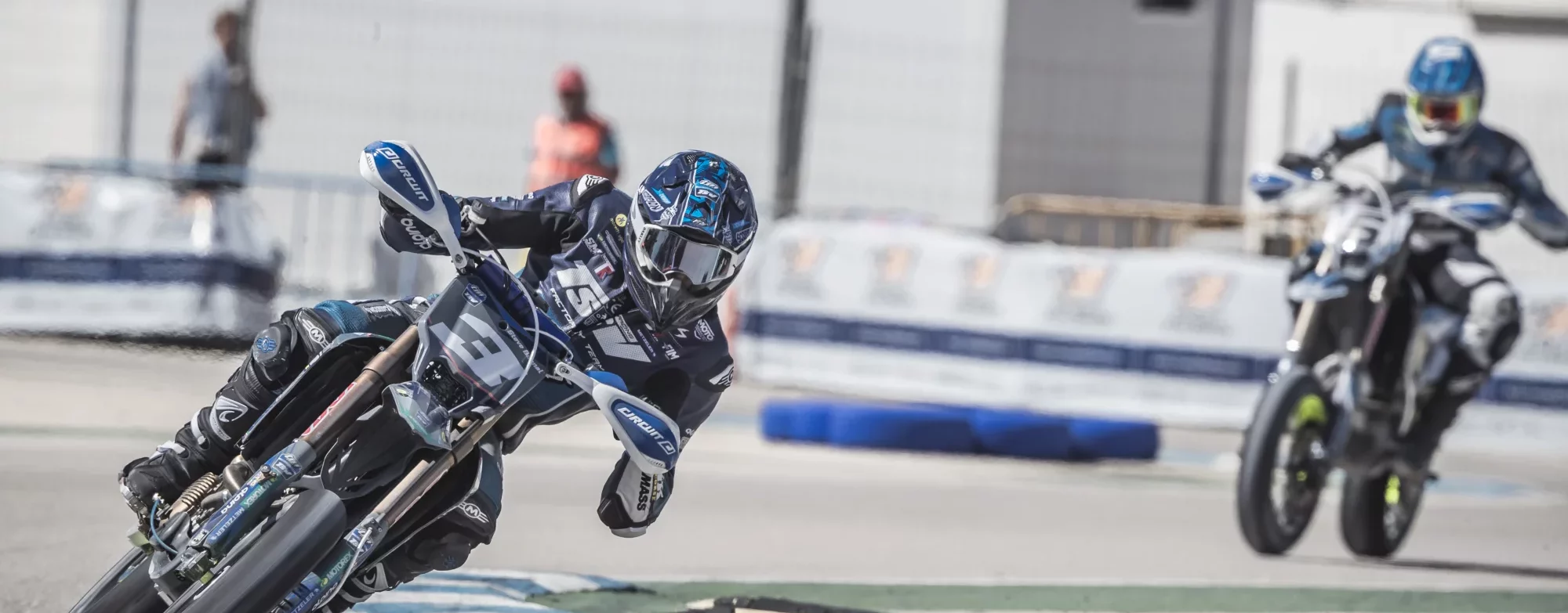 Supermoto-rijder nummer 13 stuurt scherp door een bocht op het circuit, met een tweede rijder in aantocht op de achtergrond.