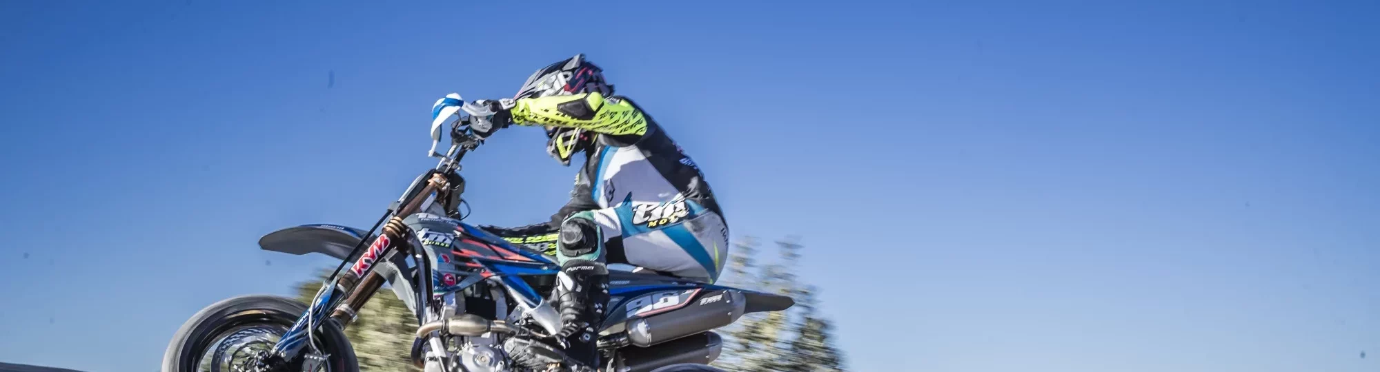 Motorcrosser in kleurrijke outfit springt met hoge snelheid over een heuvel tijdens een zonnige offroadwedstrijd.